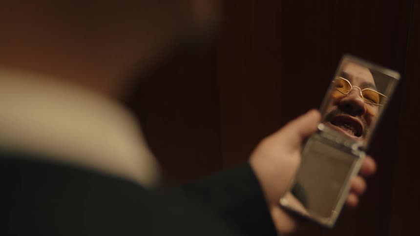 Man looks at his reflection in a small mirror inside a dimly lit room