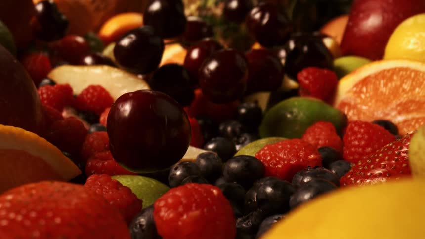 A slow motion view shows several dark red cherries and bright blue blueberries falling into a large bowl filled with a variety of colorful fruits including raspberries sliced oranges lemons and pears