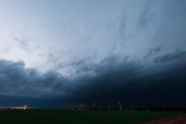 A storm cloud rolls across a green field with a few wind turbines visible on the horizon a sudden flash of lightning illuminates the sky revealing the ominous dark gray underside of the cloud