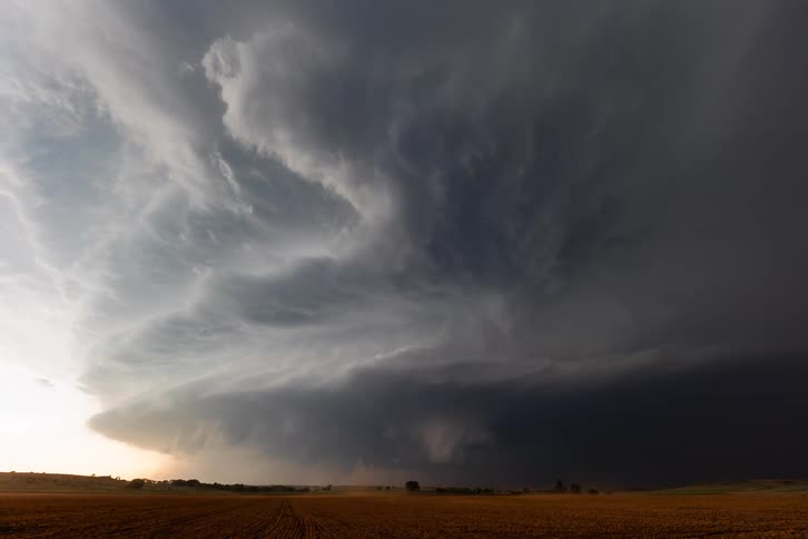 A massive dark and rapidly rotating supercell thunderstorm moving ominously across a flat open field with a wall of dust and rain being kicked up at the base of the storm