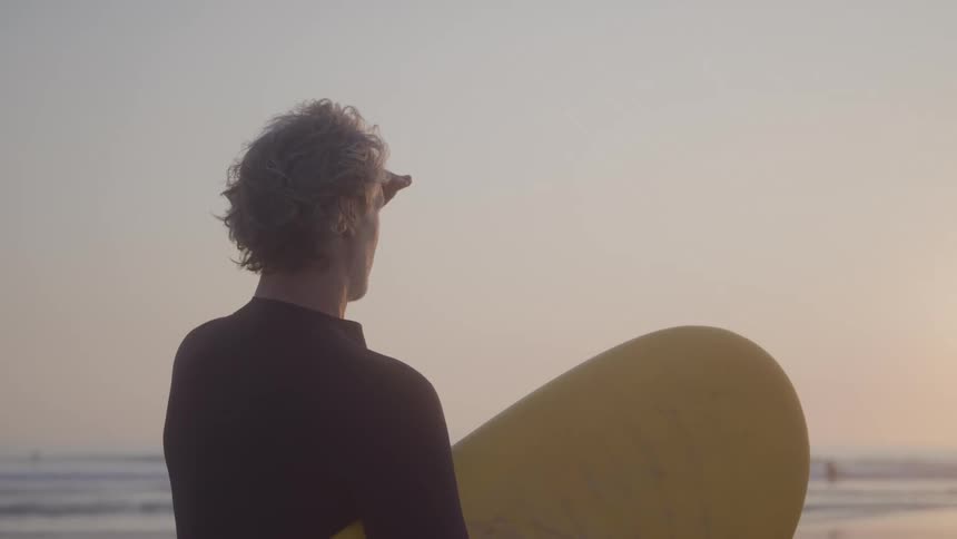 A surfer with curly grey hair wearing a black wetsuit stands on a beach holding a yellow surfboard and shades his eyes while looking out at the ocean under a pale sky