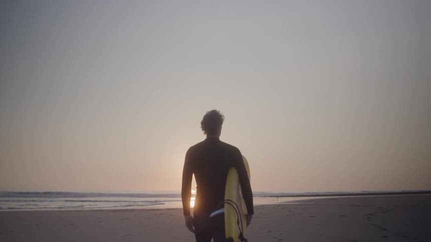 A man wearing a swimsuit holding a surfboard walking toward the ocean on the beach at sunset