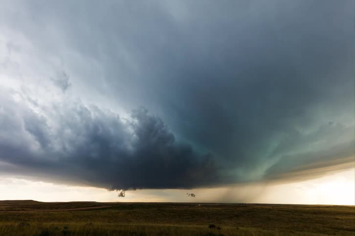 A vast dark turbulent storm cloud hangs low over a wide expanse of dry grassy land with rain visibly falling from a specific section of the cloud while the edges of the horizon remain bright