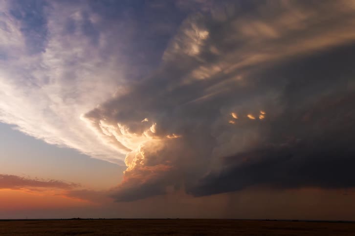 This is a dramatic timelapse of a massive storm cloud formation building and churning over a flat landscape as the sun sets the clouds are spectacularly illuminated with shifting hues of gold orange and red before darkness falls