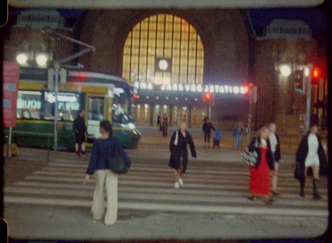 The video filmed with a retro filmlike aesthetic shows a street scene at night in front of a large train station pedestrians cross the street as a green and yellow tram passes by