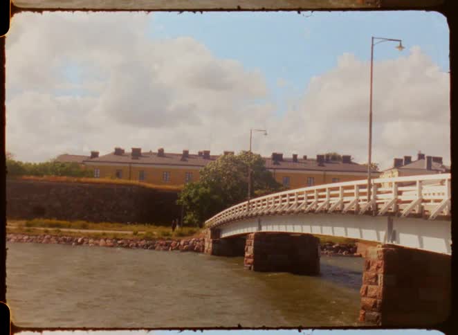 A white arched stone bridge crossing a body of water in the foreground leading to a historic fortress on the other side all under a bright blue sky with fluffy white clouds