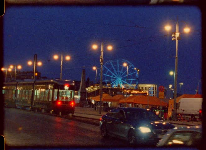 Street scene at night shows a bright ferris wheel in the distance as a tram and cars drive by with the motion of the vehicles creating streaks of light