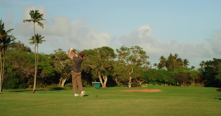A man on a golf course executes a full swing with his club hitting the golf ball off a tee on a perfectly manicured green surrounded by lush palm trees and a sandy trap in the background