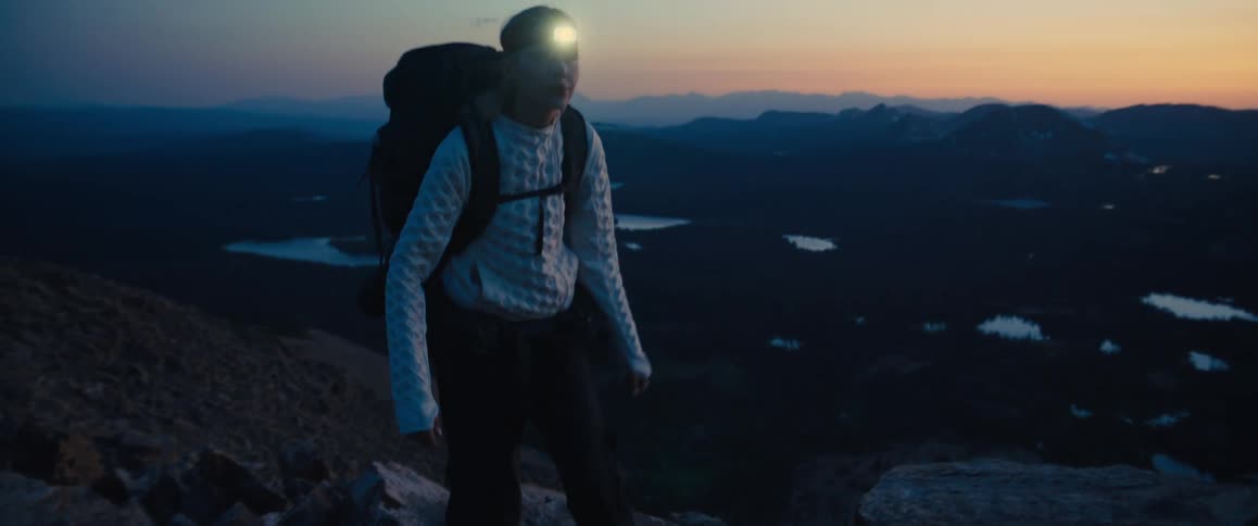 Woman wearing a backpack walks up the boulder covered slope overlooking a valley under a red tinted sky