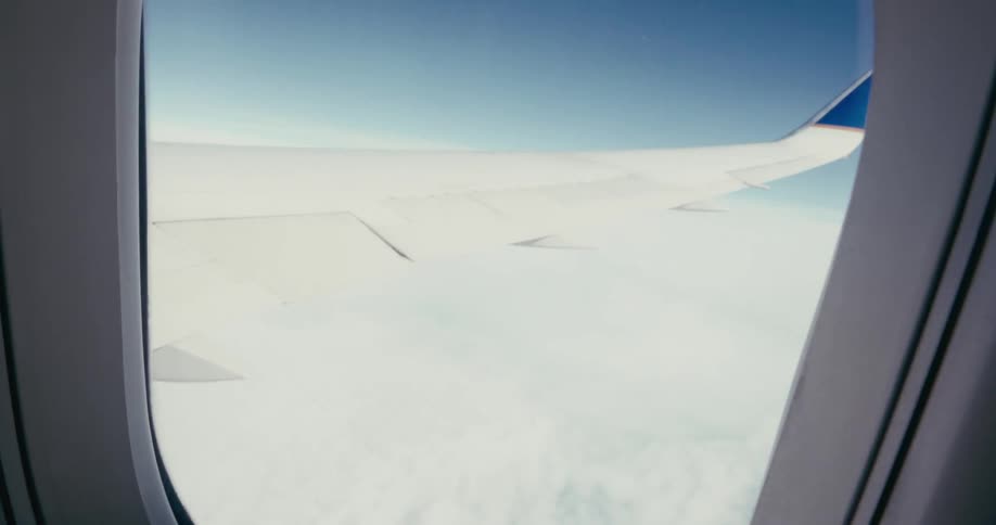 The view from an airplane window shows a white wing with a blue tip extending over a layer of clouds under a clear blue sky