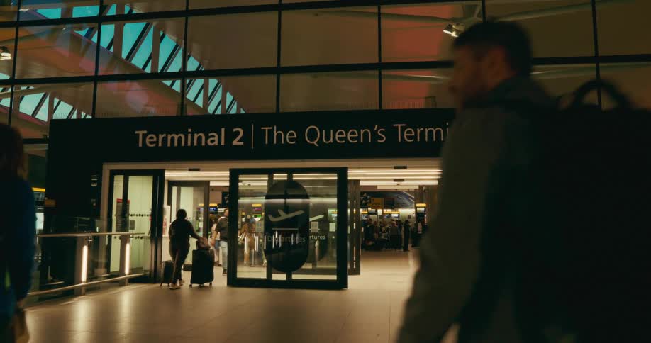 Many people of different ages and genders indoors entering and leaving a terminal in heathrow airport through sliding doors during dusk or dawn