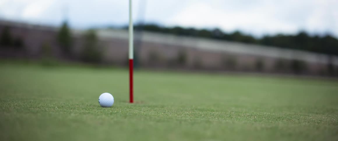 A golf course at midday with a ball positioned next to the hole marked by a flagpole while another golf ball bounces past it