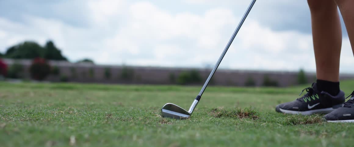 A golf club hits a golf ball on the ground on a golf course with a fence in the distance