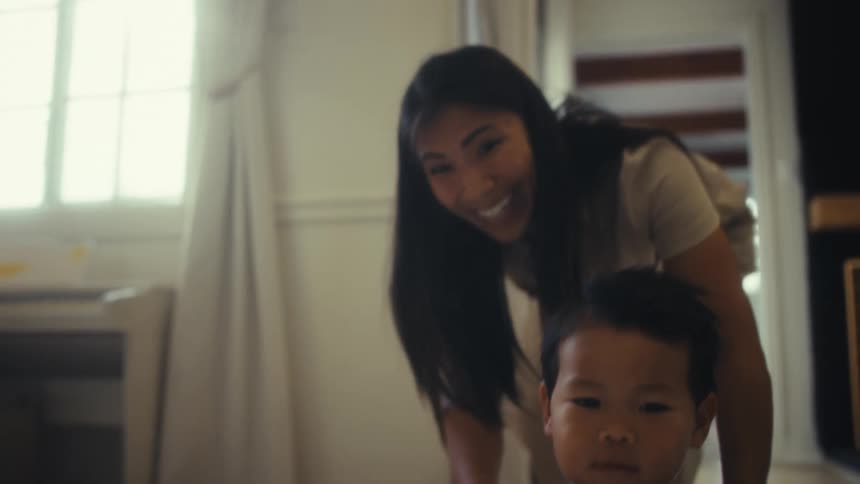A smiling mother with long dark hair pushes her young son who is sitting in a white laundry basket across a floor as he holds onto the basket and waves his hands in the air
