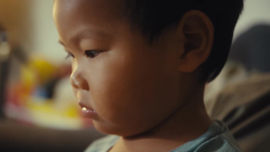 The video shows a closeup shot of a young child from the side with a pensive and focused expression on their face the childs eyes are looking down and to the side and a soft light illuminates their face from the front