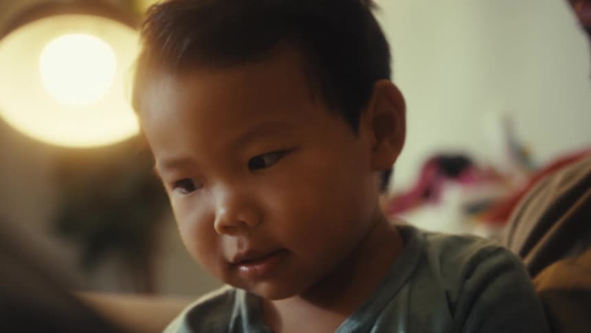 A father and his son sit together on an armchair and look at a book under a light in a indoor setting
