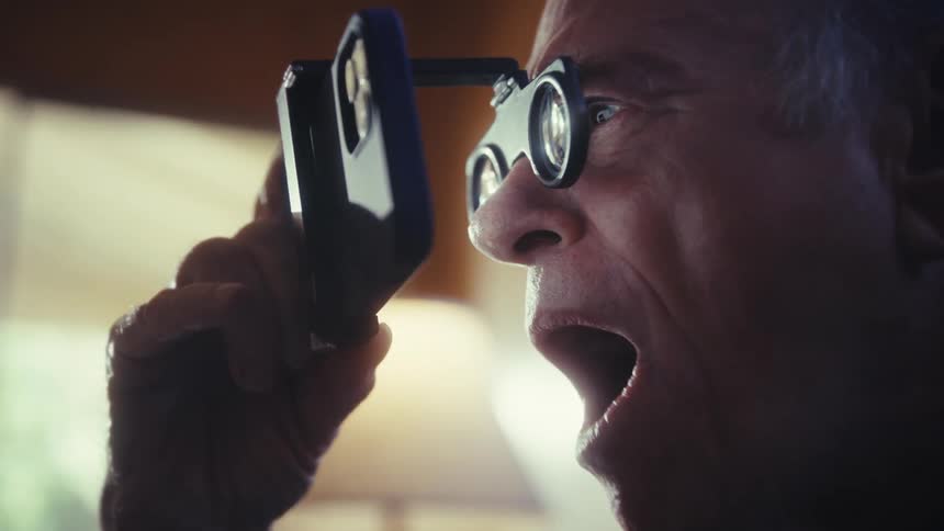 An elderly man wearing goggles holding his phone close to his eyes excitedly watching a program in his living room at midday
