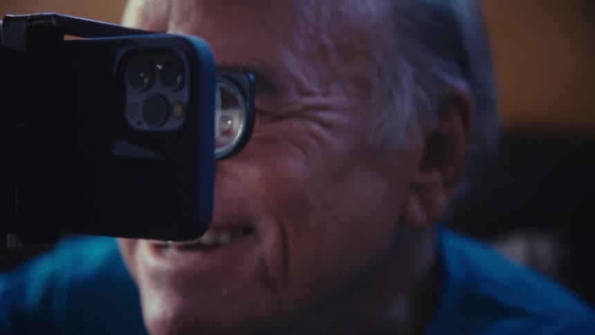 A happy person with white hair holds a phone with a small black magnifying attachment on it looking up with a big smile