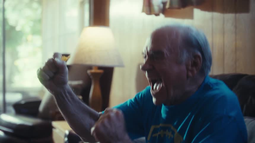 A joyful man with white hair sits on a black leather couch raising his arms in excitement as he watches something on a television out of view