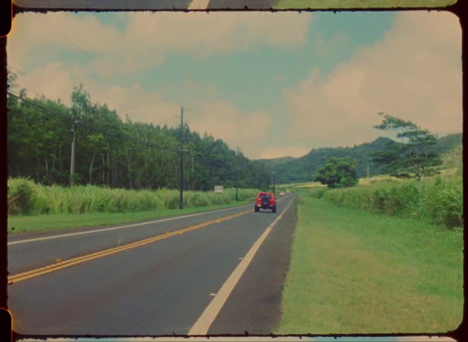 The video shows an orange jeep wrangler driving away down a twolane road the road is surrounded by lush green vegetation and trees with hills visible in the distance the footage has a vintage faded look and the video frame is bordered to resemb