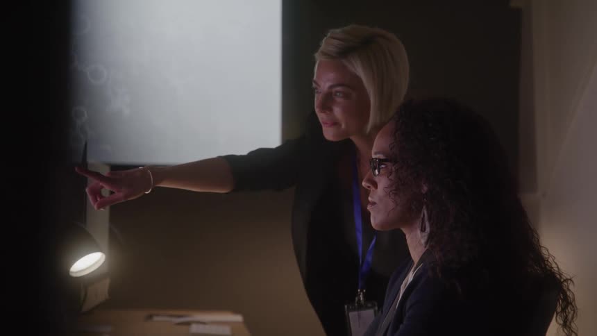 Two women are intently looking at a projected screen displaying what appears to be molecular structures or cellular patterns with one woman pointing towards the projection