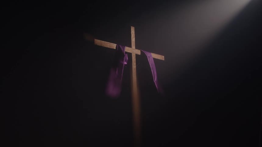 A wooden cross with a purple cloth draped over it stands in darkness illuminated by a single dramatic beam of light from above as the cloth gently sways