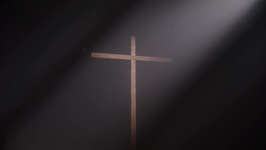 A large wooden cross stands upright in the center of the frame with beams of light shining on it from the top left against a dark foggy background