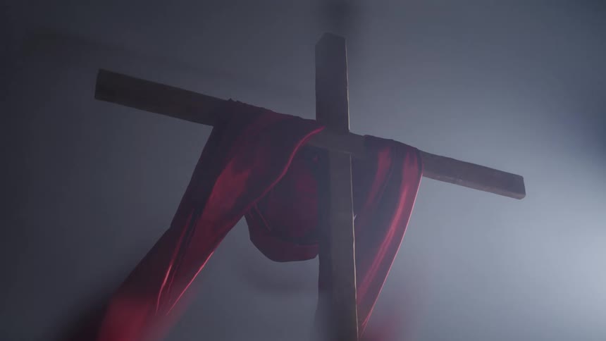 Wooden cross surrounded by smoke partially covered by a waving red cloth indoors