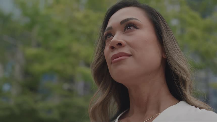 A person looks up with their face angled towards the sky with foliage and a building in the background