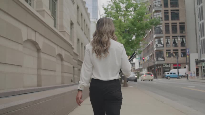 A woman with long wavy hair and a white collared shirt walks down a city sidewalk while holding a black folder or laptop