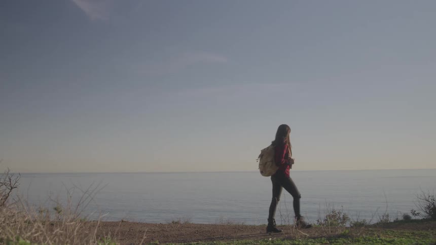 A woman with long hair and a light colored backpack walks away from the camera along a dirt path beside a vast ocean under a clear sky