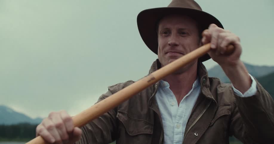 The video shows a man wearing a brown hat and jacket sitting in a boat on a body of water with mountains in the background he is holding a wooden oar the man looks up and to his left as if listening to someone or something he then looks forward again