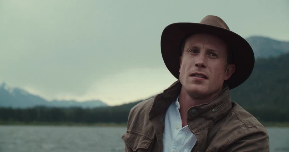 The video shows a man with a stern expression wearing a brown hat and a brown jacket over a white shirt he is outdoors on the water with mountains and trees in the background the man is holding a wooden paddle and appears to be in a small boat he loo