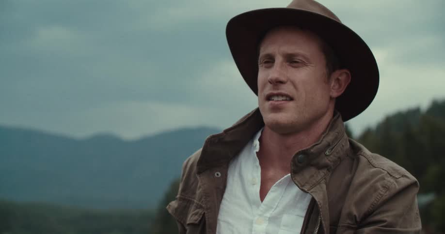 The video shows a man wearing a brown hat and jacket sitting in a boat on a body of water with mountains in the background the man is holding a wooden oar and appears to be rowing he looks off to his right and his mouth is moving as if he is speaking