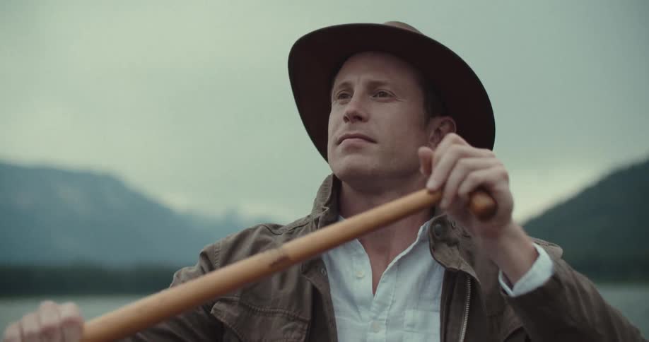 A man wearing a brown jacket and brown sun hat paddling a boat on a lake at midday