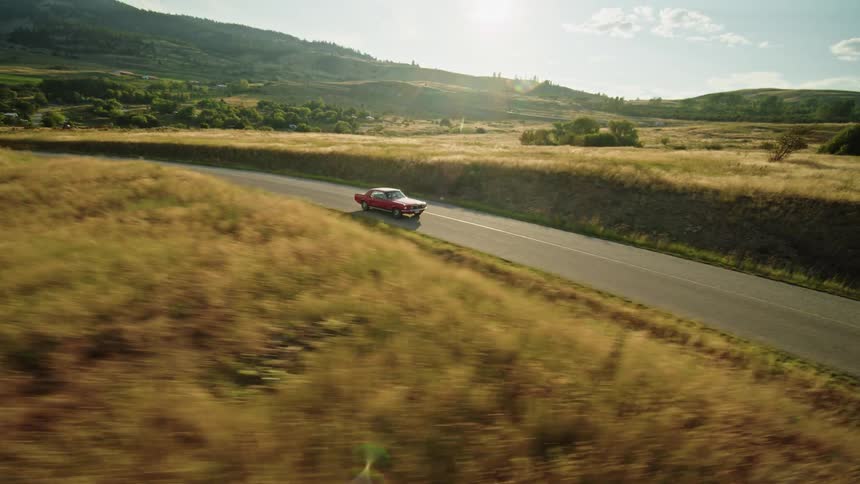 A bright red vintage car drives down a winding road through a hilly grassy landscape the sun shines brightly over the hills as the car continues its journey
