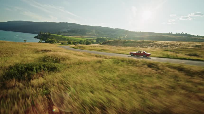 A red car drives on a winding lakeside road through grassy hills
