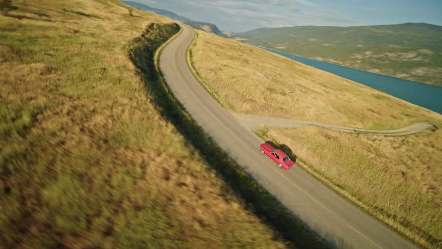 The video shows a man and a woman standing in a lush green field facing away from the camera they are looking at a picturesque landscape with mountains a lake and trees the man is wearing a hat a brown jacket and jeans while the woman is wearing a