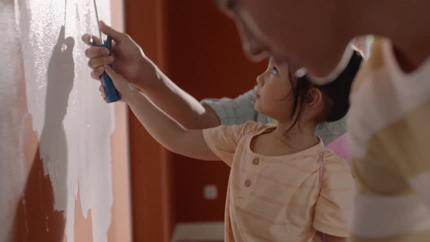 A father holding his daughters hand while painting a wall with his son painting beside them indoors at midday during a fun family moment