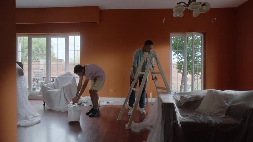 A father carries a ladder into the living room while a teenage boy walks in with a bucket of paint and both examine the bucket together