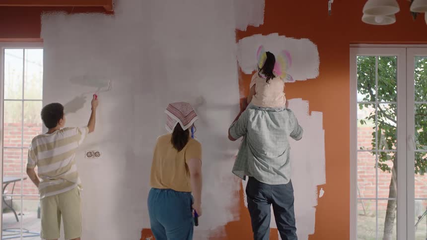 Family paints the wall together while the daughter sits on the shoulders of her father