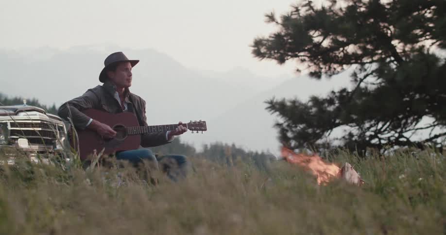 The video shows a man playing a guitar while sitting near a fire two distinct lines are present a vertical line formed by a tall tree on the right side of the screen and a horizontal line on the horizon created by the distant layered mountains