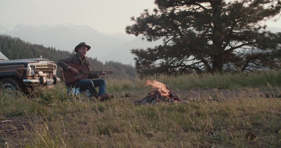 A man wearing a hat and a jacket sits on a log and plays a guitar next to a campfire with a jeep parked directly behind him and mountains in the distance