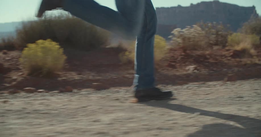 A low close view shows a person wearing blue jeans and black shoes running on a dirt road in a desert landscape