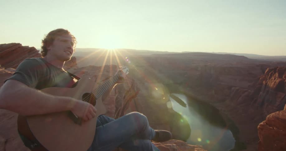 A man plays guitar while sitting on a canyon edge at sunset