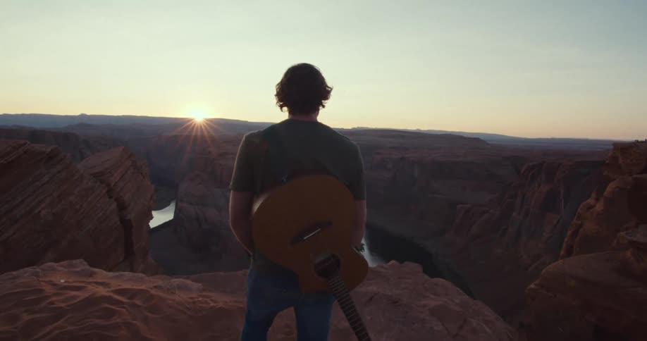 A person holding a guitaron the back gazes at a canyon and sunset