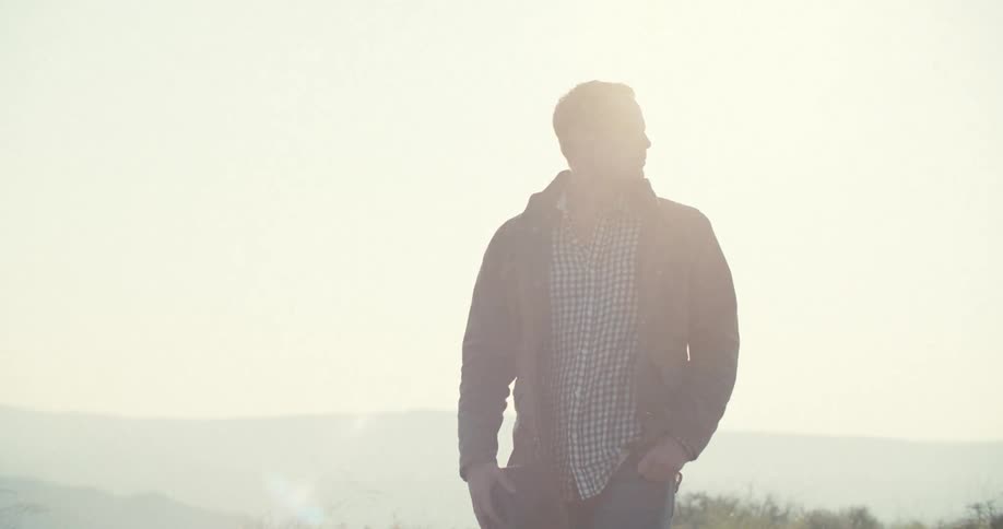 A male stands outdoors in a jacket then puts his hat on while gazing toward the mountains in the distance