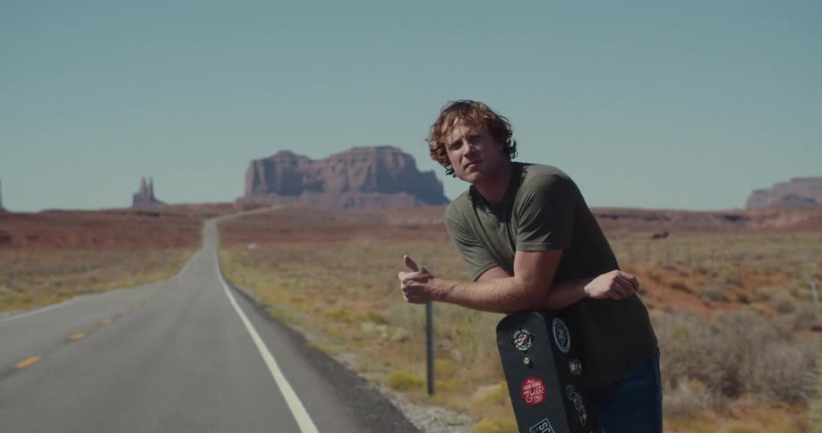 Man leaning on a guitar case on the side of the road in a desert environment with canyons in the background