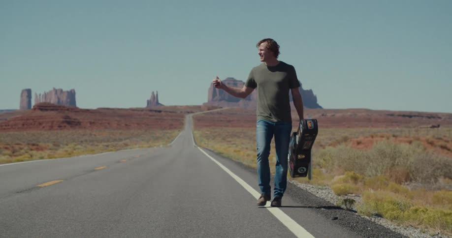 A young man holding a guitar case walks along the side of a road in the countryside