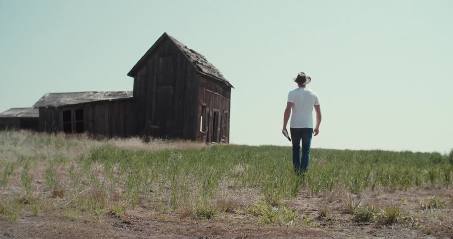 A man wearing a cowboy hat jeans and a white tshirt walks away from the camera toward an old weathered barn he is carrying an open book and walks through tall dry grass